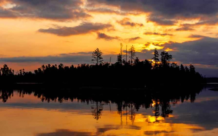 the sunset is reflected on a calm lake alongside a tree-lined shore